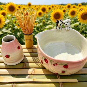 May include: A matcha tea set with a pink and white ceramic bowl decorated with strawberries, a bamboo whisk, a strawberry-decorated ceramic container, and a bamboo scoop. Two small figurines of a cat and a dog are perched on the bowl's rim. The set is on a bamboo surface.