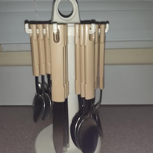 Könnte beinhalten: Ein weißer Plastik-Besteckhalter mit einem runden Griff oben. Der Halter hat Schlitze für Löffel, Gabeln und Messer. Das Besteck ist aus Edelstahl und hat beigefarbene Kunststoffgriffe.