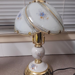 May include: A vintage table lamp with a white and gold finish. The lamp features a floral patterned lampshade and base, with gold accents. The lamp is approximately 46 cm tall.