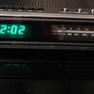 May include: A black Sunbeam electronic alarm clock radio with a digital green display showing 2:02 PM. The device features a horizontal analogue FM and AM radio frequency tuner, top-mounted control buttons, and a silver trim accenting the front panel.