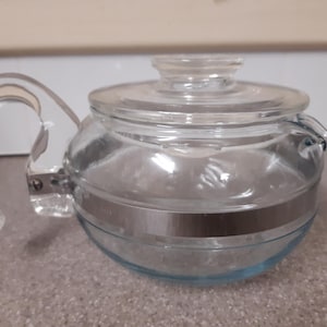 May include: A clear glass teapot with a curved, transparent handle and a matching lid. The teapot has a silver band around the middle and a slight blue tint at the base. The spout is small and curved.