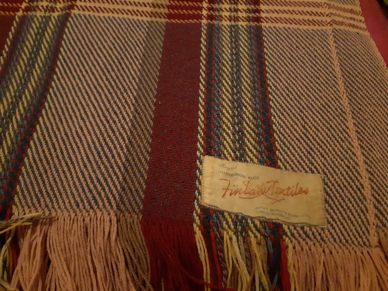 Peut inclure: Une couverture tiss&eacute;e avec un motif &agrave; carreaux dans les tons de rouge, bleu, jaune et beige. La couverture a un bord &agrave; franges et une &eacute;tiquette qui indique "Finlay Textiles".