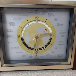 Könnte beinhalten: Eine Vintage Rhythm Quartz Weltzeituhr mit einem braunen Holzrahmen und einem goldfarbenen Zifferblatt. Die Uhr zeigt die Zeit in verschiedenen Städten der Welt an. Das Zifferblatt hat eine Weltkarte in der Mitte.