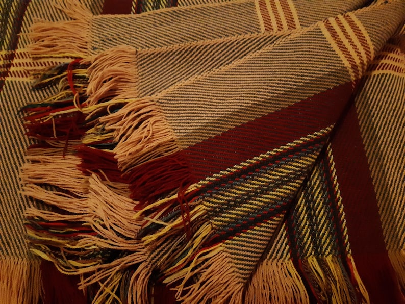 Peut inclure: Une couverture pli&eacute;e avec un motif tiss&eacute; de rayures altern&eacute;es dans les tons beige, bordeaux et gris. La couverture a un bord &agrave; franges de couleur beige clair. La texture g&eacute;n&eacute;rale semble douce et accueillante.