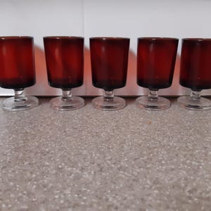 Puede incluir: Conjunto de seis vasos de chupito de vidrio rojo con tallos transparentes. Los vasos están dispuestos en fila sobre una superficie marrón claro.