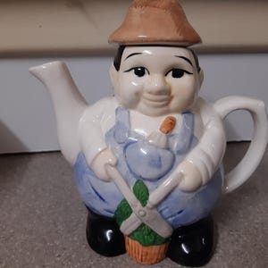 Könnte beinhalten: Eine Keramik-Teekanne in Form eines Gärtners, mit braunem Hut, weißem Hemd und blauer Latzhose. Die Teekanne hat einen weißen Ausguss und Henkel, und der Gärtner hält eine Schere und eine Pflanze. Der Sockel ist schwarz.