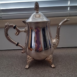 May include: A silver teapot with a decorative finial on the lid and three ornate feet. The teapot has a black handle and spout.
