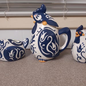 May include: A ceramic tea set with a rooster theme, in white and navy blue. The set includes a teapot with a handle and lid, and two hen-shaped containers. The rooster and hen designs feature intricate patterns and orange accents.