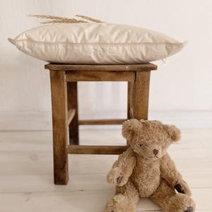 Puede incluir: Una almohada beige con una espiga de trigo decorativa descansa sobre un pequeño taburete de madera. Un oso de peluche marrón está sentado en el suelo frente al taburete. La escena se desarrolla contra una pared blanca y un suelo de madera clara.