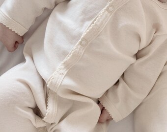 May include: A cream-colored baby onesie with long sleeves and a scalloped trim. The onesie has a button closure down the front.