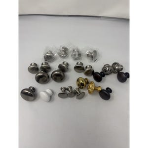 Peut inclure: Une collection de boutons de placard variés dans différentes finitions, notamment en nickel brossé, laiton poli, noir mat et céramique blanche. L'assortiment présente des designs ronds, lisses et texturés, certains étant emballés individuellement.