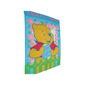 May include: A rectangular Easter-themed banner featuring Winnie the Pooh holding a decorated Easter egg. The banner has a light blue background with pink and yellow flowers and a green picket fence. The text "EASTER IS COMING" is at the top.