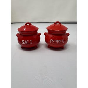 May include: A pair of red ceramic salt and pepper shakers. Each shaker is a small, round pot with a lid and small handles. The word "SALT" is printed on one shaker, and "PEPPER" on the other.