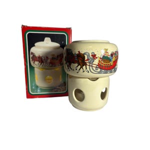 May include: A cream-colored Christmas candle warmer with a Santa Claus sleigh design. The warmer has a round base with two openings and a top piece with a lid. The box shows a similar design. Festive holiday decor.