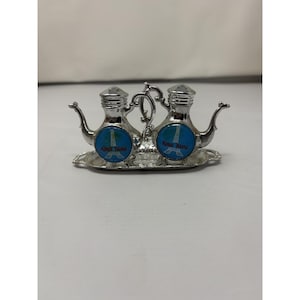 May include: Silver-toned salt and pepper shaker set. Each shaker has a teapot-like design with a blue and white circular emblem featuring a tower and the text "Kings Island". The set sits on a matching tray.