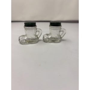 May include: Two clear glass boot-shaped salt and pepper shakers with dark green lids. Each shaker has a handle and is designed to resemble a small boot. The shakers are displayed on a white surface.