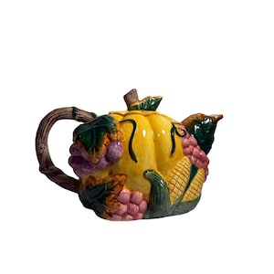 May include: A ceramic teapot shaped like a yellow pumpkin, adorned with green leaves, purple grapes, and corn. The handle is brown, resembling a tree branch. The spout is on the right side, and the lid is at the top.
