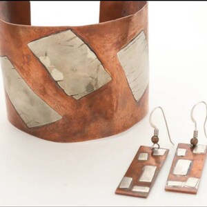 Op de afbeelding: Een koperen manchetarmband en bijpassende oorbellen set. De armband en oorbellen hebben rechthoekige, zilverkleurige metalen accenten. De oorbellen hebben een haaksluiting. De sieradenset wordt op een witte achtergrond weergegeven.