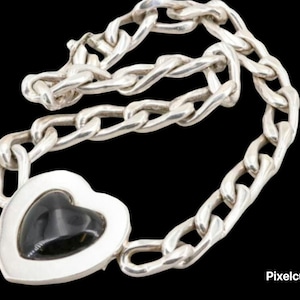Op de afbeelding: Een zilveren kettingarmband met een hartvormige hanger. De hanger heeft een zwarte steen in een zilveren frame. De schakels van de armband zijn dik en getextureerd, wat een gedurfd ontwerp creëert. De armband wordt tegen een zwarte achtergrond getoond.