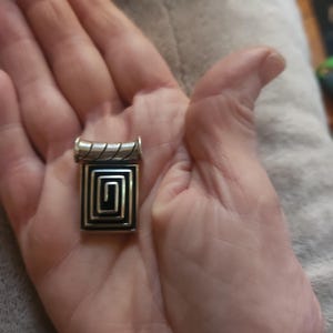 Puede incluir: Un colgante plateado con un dise&ntilde;o geom&eacute;trico negro y plateado. El colgante tiene forma rectangular con un patr&oacute;n en espiral en el centro. La parte superior del colgante tiene un dise&ntilde;o cil&iacute;ndrico. El colgante se sostiene en una mano.