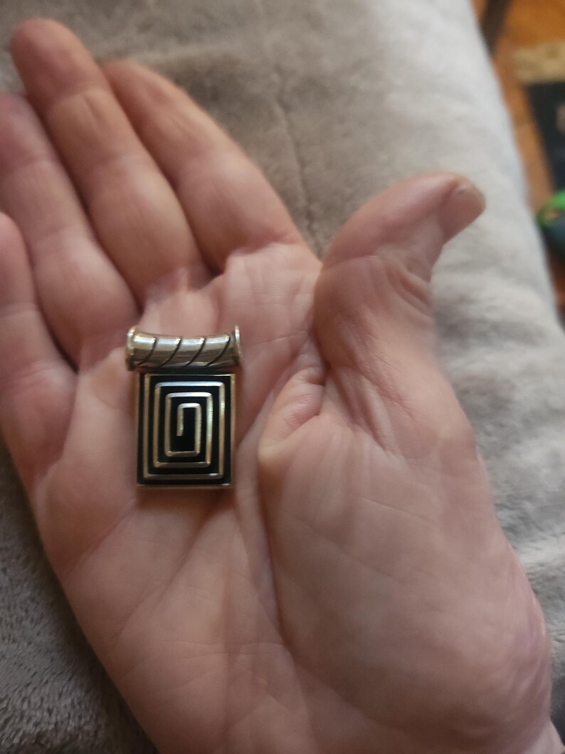Puede incluir: Un colgante plateado con un dise&ntilde;o geom&eacute;trico negro y plateado. El colgante tiene forma rectangular con un patr&oacute;n en espiral en el centro. La parte superior del colgante tiene un dise&ntilde;o cil&iacute;ndrico. El colgante se sostiene en una mano.