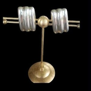 May include: A pair of silver-toned hoop earrings with a ribbed, multi-band design displayed on a gold-toned metal stand. The earrings feature a curved, rectangular shape with three distinct horizontal ridges running across the surface.