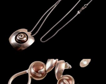 Puede incluir: Un collar con colgante plateado con un colgante cuadrado con un diseño en espiral. El collar tiene una cadena delicada y un par de pendientes a juego con un diseño en espiral similar. La joyería está sobre un fondo negro.