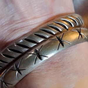 May include: Two silver-toned metal bangles. The top bangle features a series of diagonal etched lines. The bottom bangle displays a repeating pattern of four-pointed star-shaped engravings. Both pieces have a smooth, polished metallic finish.