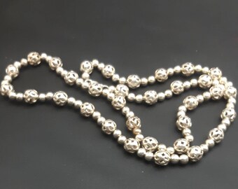 May include: A silver necklace featuring a combination of small, solid beads and larger, openwork beads. The necklace is displayed against a dark backdrop, highlighting its detailed design and metallic lustre. The beads are uniformly spaced.