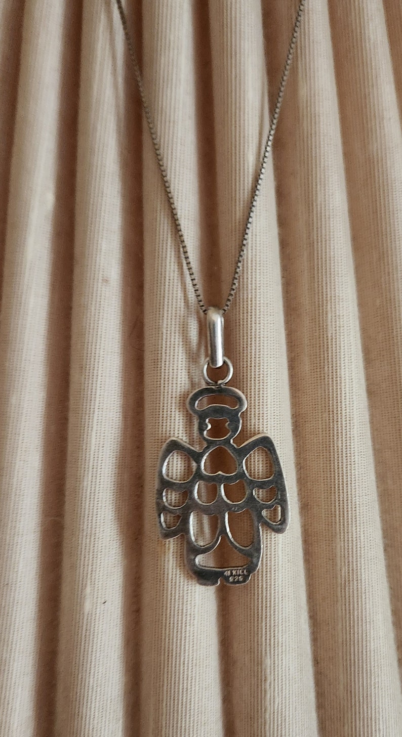 Puede incluir: Collar con colgante de plata con dise&ntilde;o de &aacute;ngel. El colgante, una silueta calada de &aacute;ngel con halo, cuelga de una fina cadena. El fondo es de tela beige. Marcado con '4 KICK 925'.