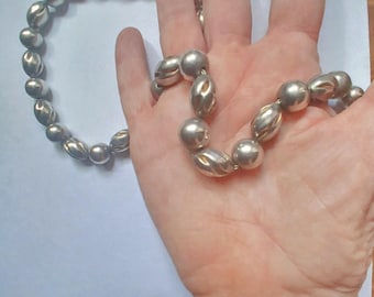 May include: A silver necklace with alternating round and oval beads. The necklace is shown against a light background, highlighting its polished, reflective surface. The beads have a smooth, metallic finish, reflecting light.