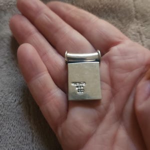 Puede incluir: Un colgante rectangular de plata con una parte superior redondeada y una base rectangular. El colgante se sostiene en una mano. El texto "MEXICO" y otras marcas son visibles en la base.