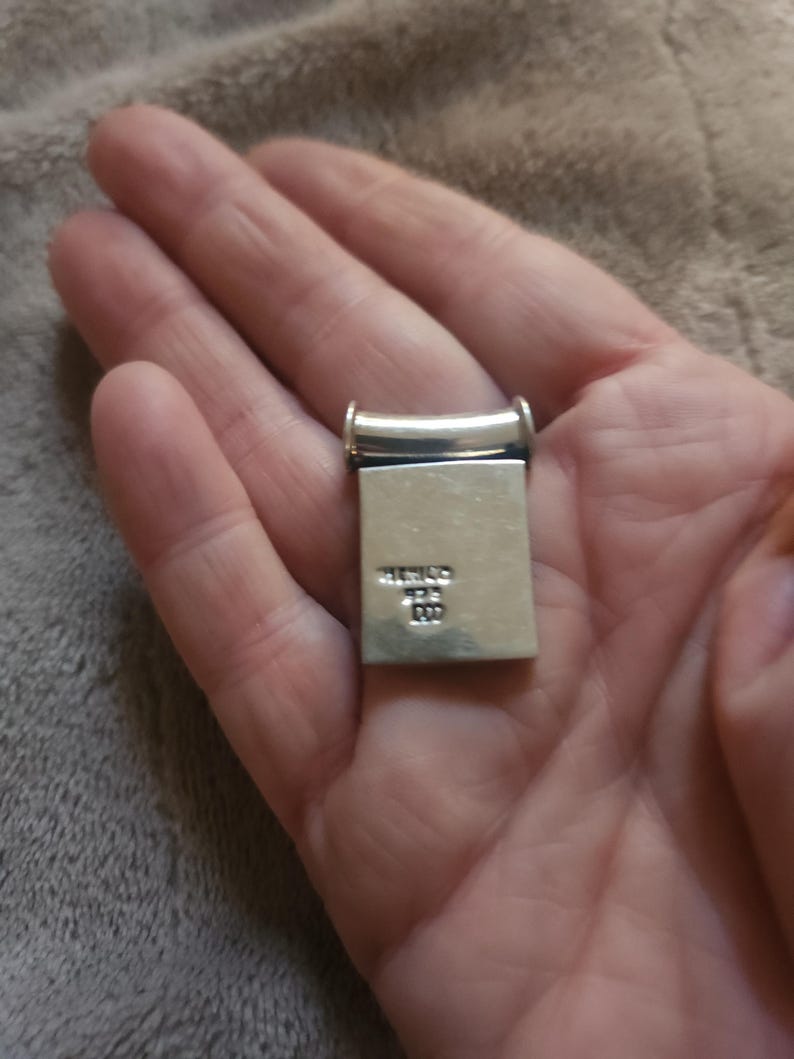 Puede incluir: Un colgante rectangular de plata con una parte superior redondeada y una base rectangular. El colgante se sostiene en una mano. El texto "MEXICO" y otras marcas son visibles en la base.