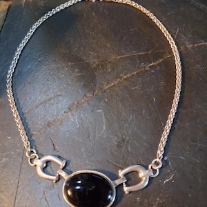 May include: A silver-toned necklace featuring a braided chain and a central oval-shaped black stone pendant. The pendant is flanked by two metallic horseshoe-shaped links that connect the stone setting to the chain, secured with a lobster claw clasp.