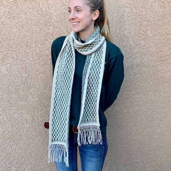 May include: A white and green knitted scarf with fringe. The scarf is worn by a person with long brown hair and a green shirt.