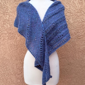 May include: A blue knitted shawl with a textured design is displayed on a mannequin. The shawl features a V-neck shape and a draped design. The knitted fabric has a speckled appearance and a decorative openwork pattern along the edges.