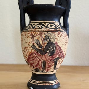 May include: A black and beige ceramic vase with two handles, featuring a black-figure style illustration of a bearded person playing a lyre. The vessel has a wide rim, a tapered body, and a circular base, decorated with a wave pattern near the top.