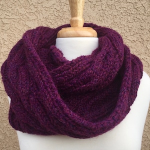 May include: A deep purple infinity scarf knitted in a textured pattern. The scarf is soft and warm, perfect for cold weather.