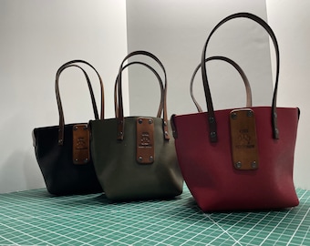 May include: Three leather tote bags with brown leather handles. The bags are black, green, and burgundy. Each bag has a brown leather patch with the text "The Fox Den" embossed on it.