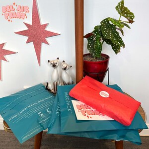 May include: An image featuring teal and red packaging, with the text "BEL AIR PRINT" visible. The scene includes decorative starbursts, ceramic cat figurines, and a potted plant. The packaging is labeled "HEROPACK" and "HOME-COMPOSTABLE."