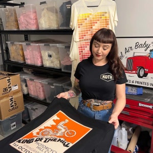 May include: A woman holding a black t-shirt with an orange and white graphic. The graphic reads "KING OF THE HILL MOTORCYCLE HILL CLUB 1st OCTOBER SPRINGRANG". A black t-shirt with "KOTH" logo is also visible.