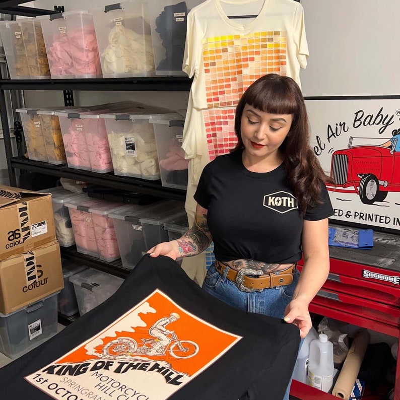 May include: A woman holding a black t-shirt with an orange and white graphic. The graphic reads "KING OF THE HILL MOTORCYCLE HILL CLUB 1st OCTOBER SPRINGRANG". A black t-shirt with "KOTH" logo is also visible.