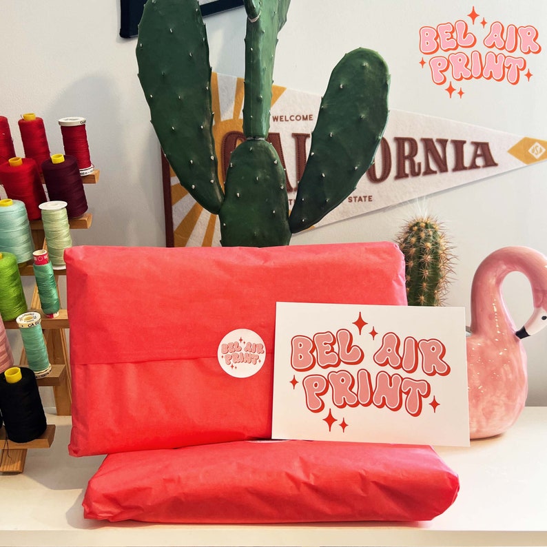 May include: Two packages wrapped in bright coral tissue paper, with a sticker and a card that read "BEL AIR PRINT" in pink and white. A pink flamingo statue and a cactus are also visible.