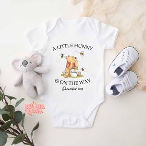 May include: White baby onesie with the text "A Little Hunny Is On The Way December 2025" and a Winnie the Pooh graphic. Includes a gray koala plush toy and a pair of white baby sneakers.