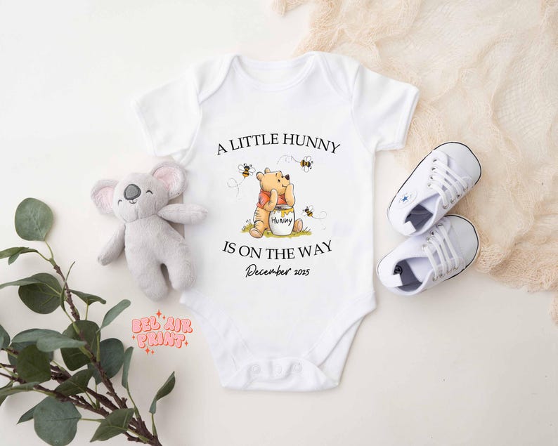 May include: White baby onesie with the text "A Little Hunny Is On The Way December 2025" and a Winnie the Pooh graphic. Includes a gray koala plush toy and a pair of white baby sneakers.