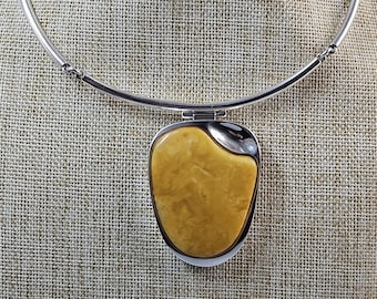 May include: A silver-toned necklace with a large, pendant. The pendant features a light yellow, oval-shaped stone set in a silver frame. The necklace is displayed on a mannequin.