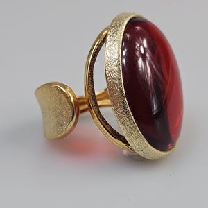 Könnte beinhalten: Ein goldfarbener Ring mit einem großen, ovalen, tiefroten Edelstein. Der Edelstein ist in einem strukturierten Goldband gefasst, mit einem glatten, halbmondförmigen Akzent an der Seite. Der Ring hat ein modernes, elegantes Design.