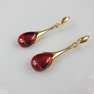 Può includere: Un paio di orecchini pendenti dorati. Ogni orecchino presenta una pietra rossa traslucida a forma di goccia, incastonata in una montatura in metallo dorato. Design semplice ed elegante.