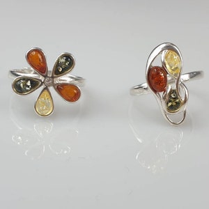 May include: Two silver rings set with amber stones. The first ring features a flower design with five amber stones in shades of green, yellow, and orange. The second ring has a more abstract design with three amber stones in shades of yellow, orange, and green.