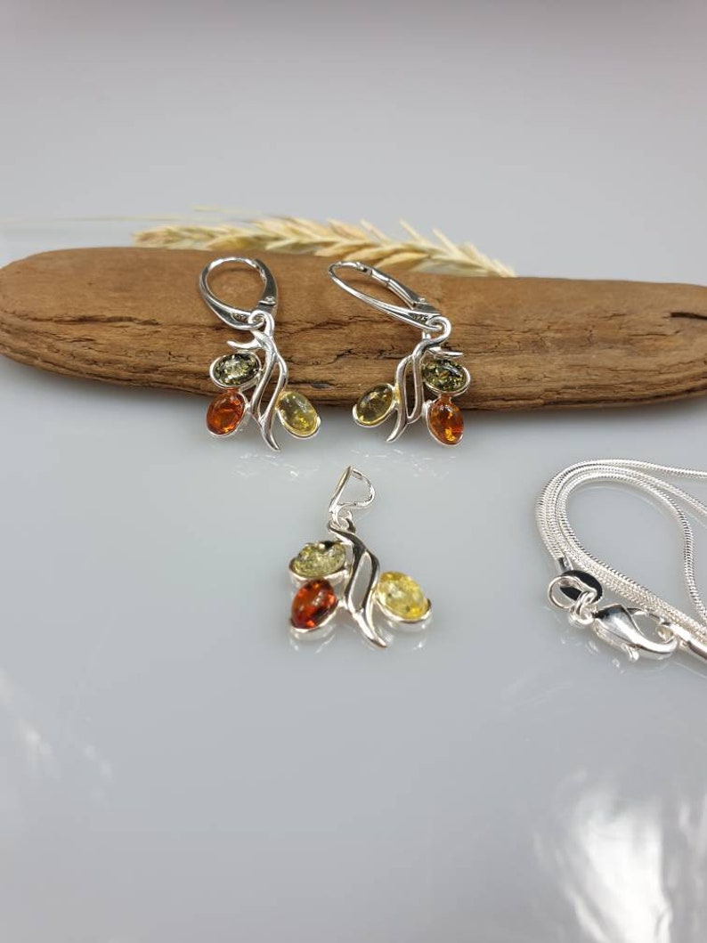Op de afbeelding: Een zilveren ketting- en oorstekerset met een hanger en hangende oorbellen met drie amberstenen in tinten groen, geel en oranje.